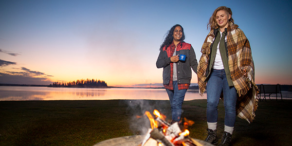 Two women smile around a campfire during a sunset at Astotin Lake in Elk Island National Park.