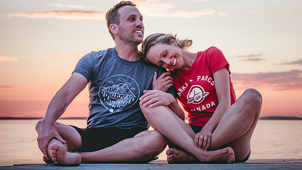 A man and a woman wearing Parks Canada t-shirts sit on a dock by a lake as the sun sets.